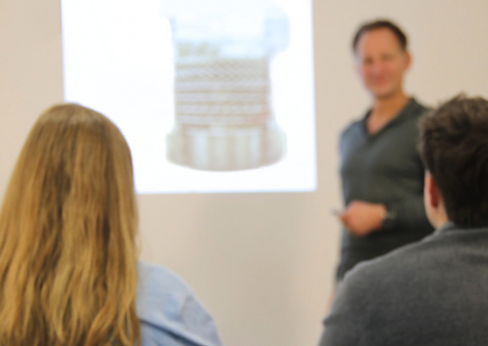 Zwei Personen hören bei einer Schulung zu. Es spricht ein Mann vor einer auf die Wand projizierten Präsentation.